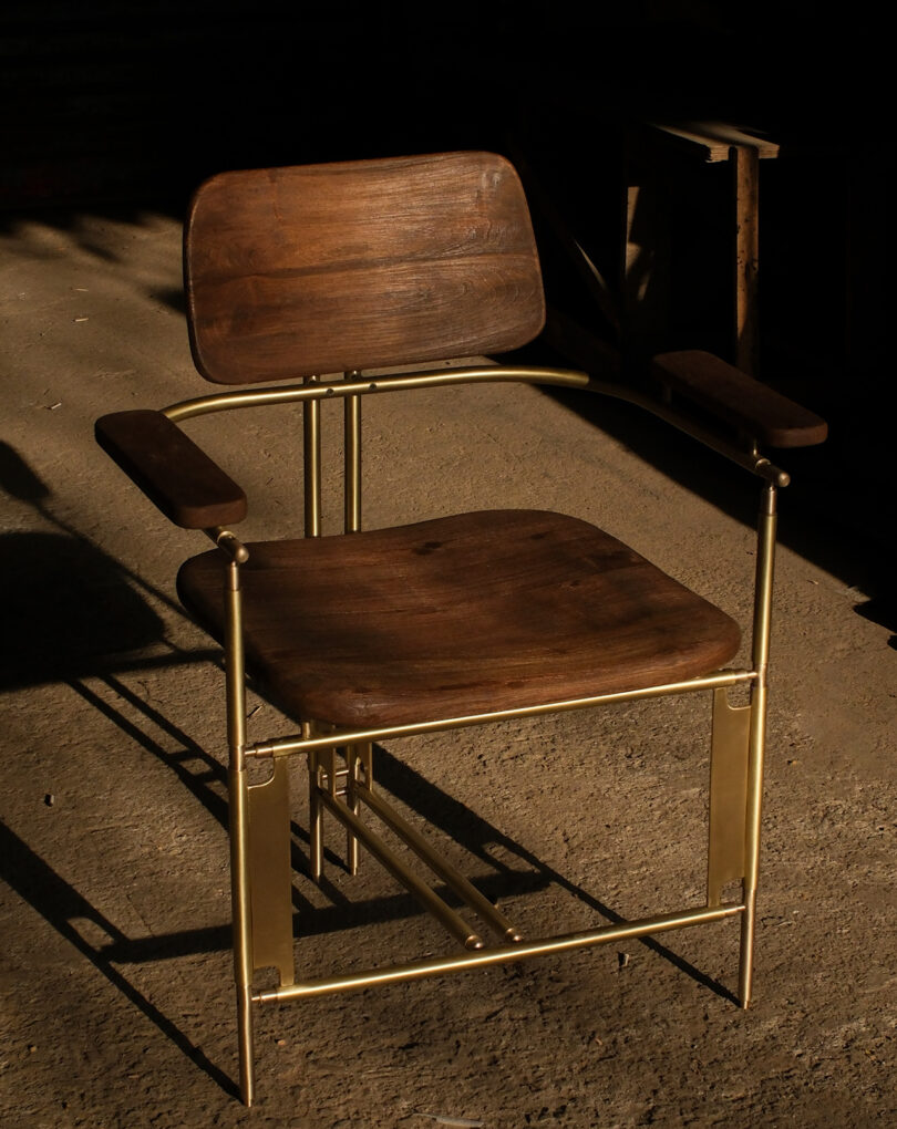 A wooden chair with a brass metal frame and armrests sits on a concrete floor in partial sunlight.