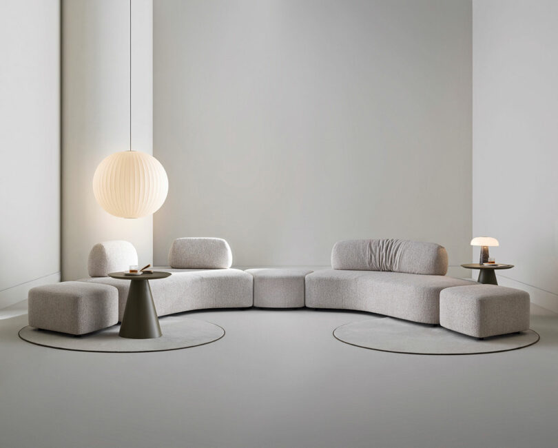 A modern living room with a light gray sectional sofa, two round side tables, a large hanging lamp, a small table lamp, and minimal decor on a light gray floor and walls.