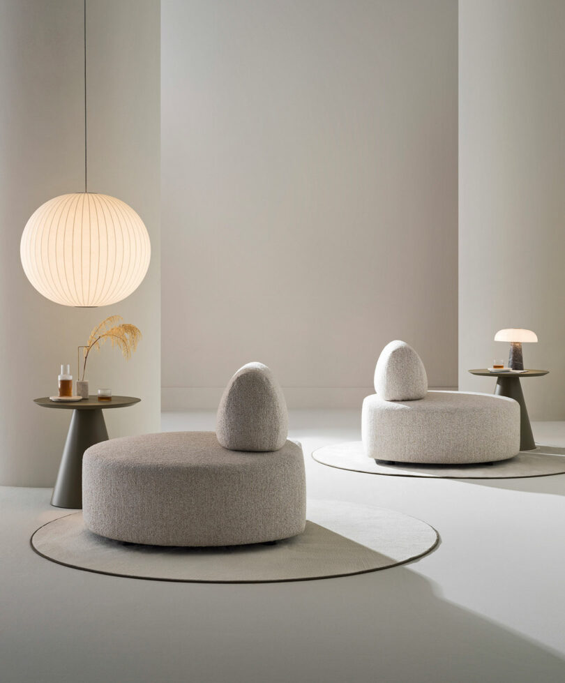 Two modern round lounge chairs with rounded backrests sit on circular rugs, accompanied by small side tables and minimalist lamps in a softly lit, neutral-toned room.