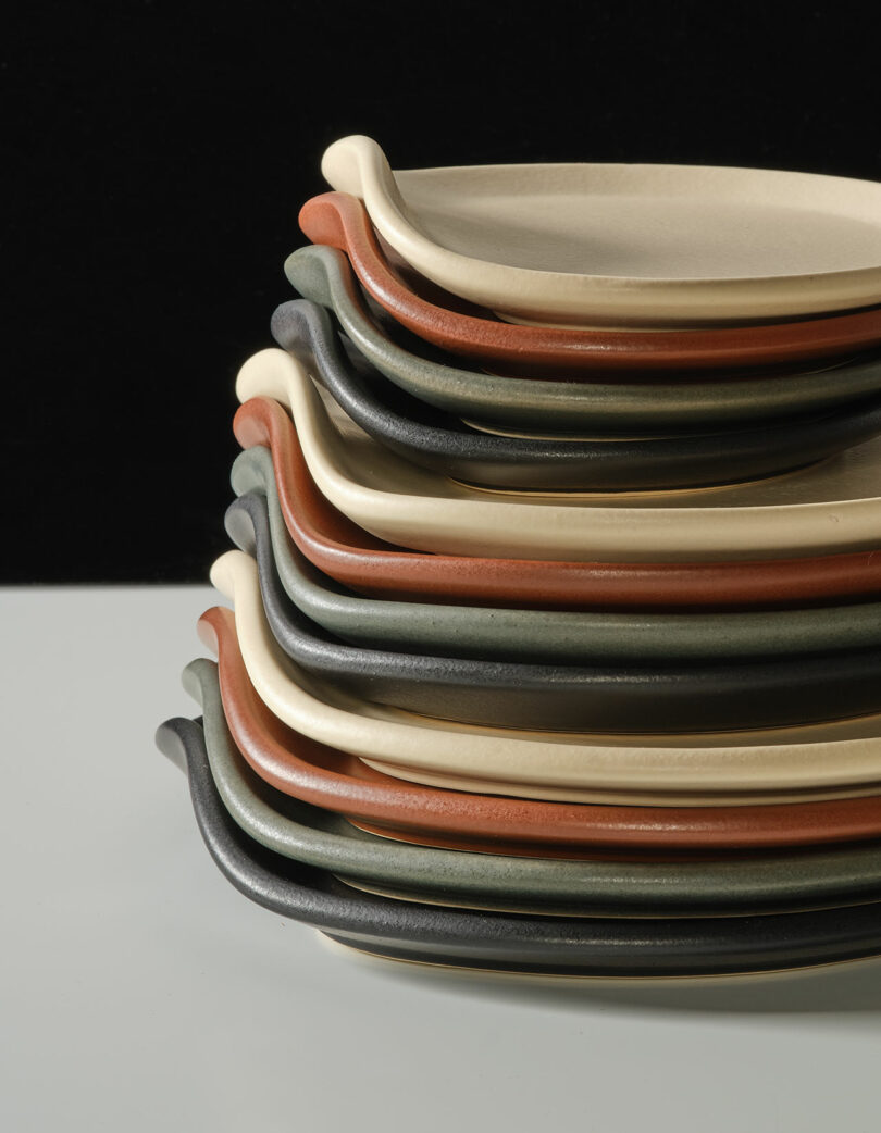 A stack of oval ceramic plates in shades of beige, black, green, and rust, arranged neatly against a plain background.