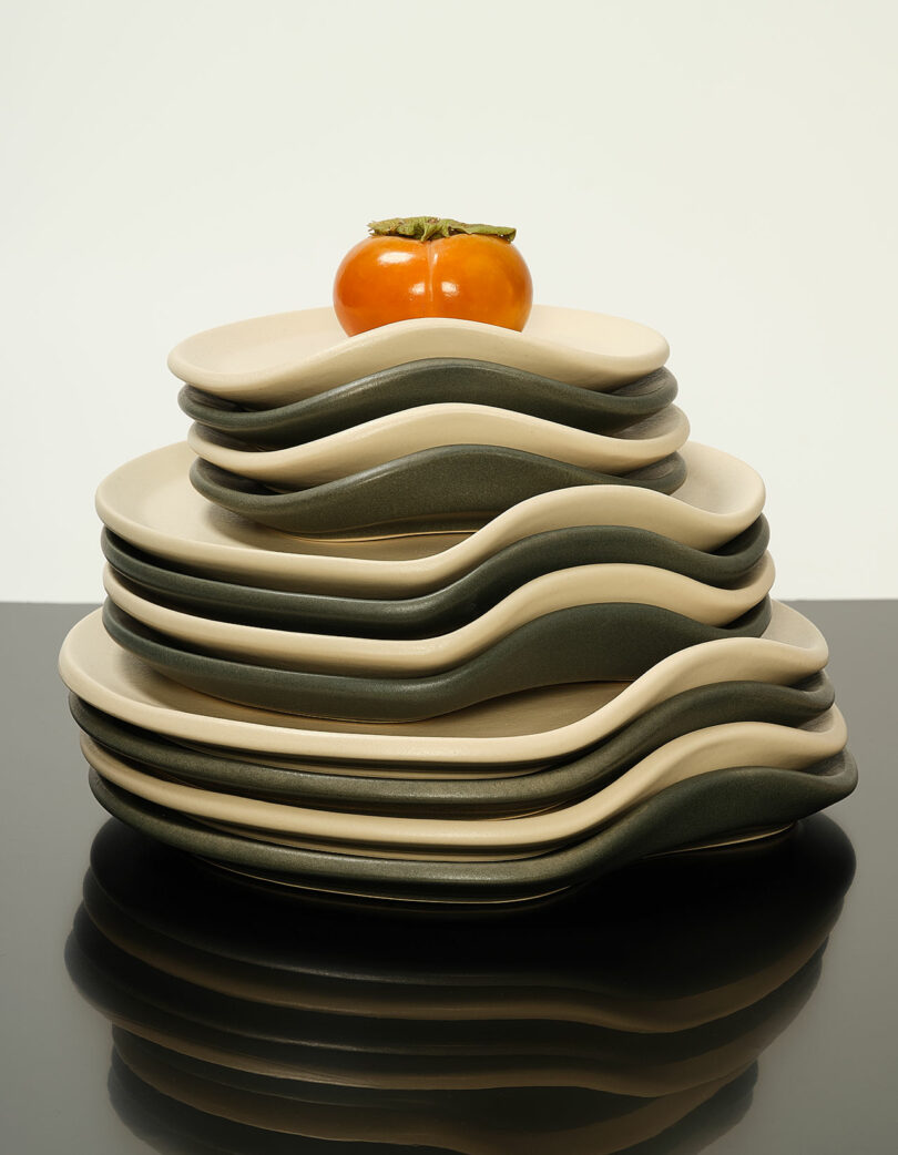 A stack of wavy-edged plates in beige and dark green tones with a single persimmon placed on the top plate.