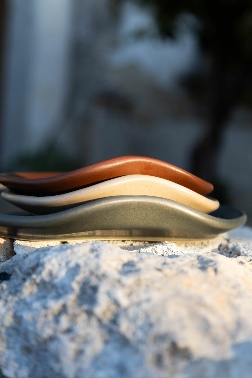 Three wavy ceramic plates in brown, beige, and gray are stacked on a rough stone surface, with a blurred outdoor background.