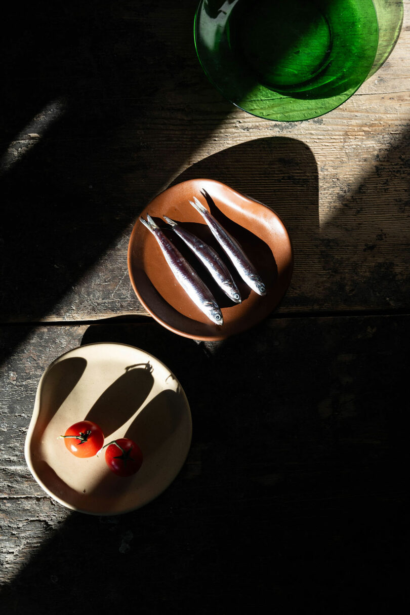 Three small fish on a brown plate and two tomatoes on a beige plate are placed on a rustic wooden table with strong, diagonal sunlight and shadows. A green glass bowl is partially visible at the top.