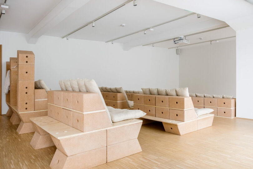 A minimalist room with modular, light brown block seating arranged in rows, each topped with white cushions, and a projector mounted on the ceiling