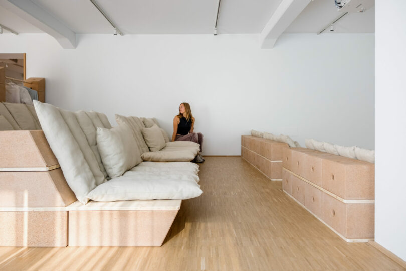 A woman sits alone on a large, cushioned wooden bench in a minimalist room with white walls and a wooden floor