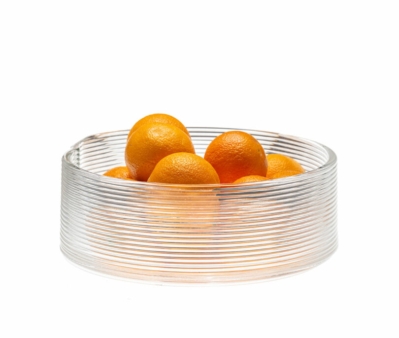 A clear, ribbed glass bowl containing several whole oranges, set against a plain white background.