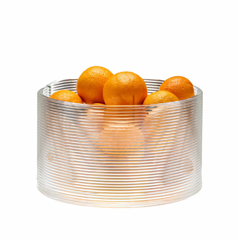 A clear, ribbed glass bowl filled with several whole oranges, photographed against a white background.