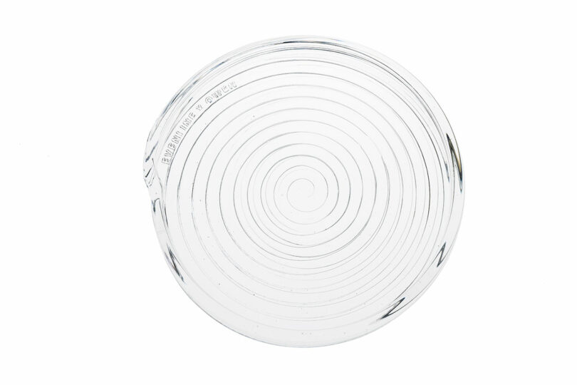A clear round glass object with concentric circular ridges, resembling a lens or a textured coaster, photographed on a white background.