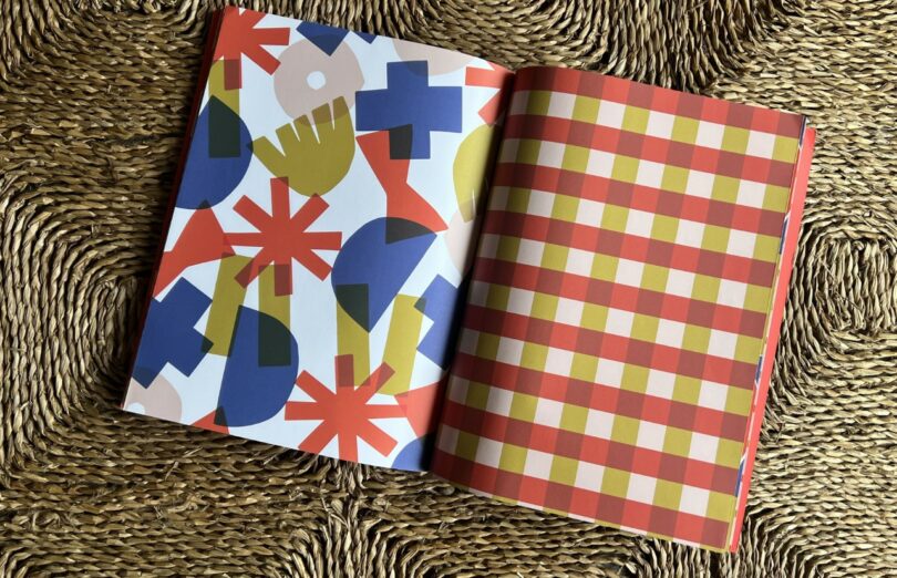 An open book, featured in Design Milk Take 5, displays abstract, multicolored geometric shapes on the left page and a red, yellow, and white checkered pattern on the right page, resting on a textured woven surface