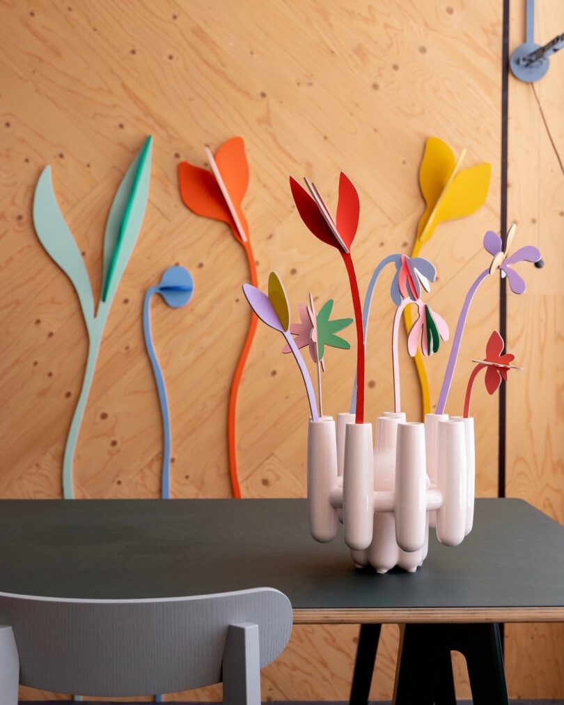 A modern room with a black table, gray chair, white ceramic vase holding colorful abstract flowers, and stylized flower decorations on a wooden wall