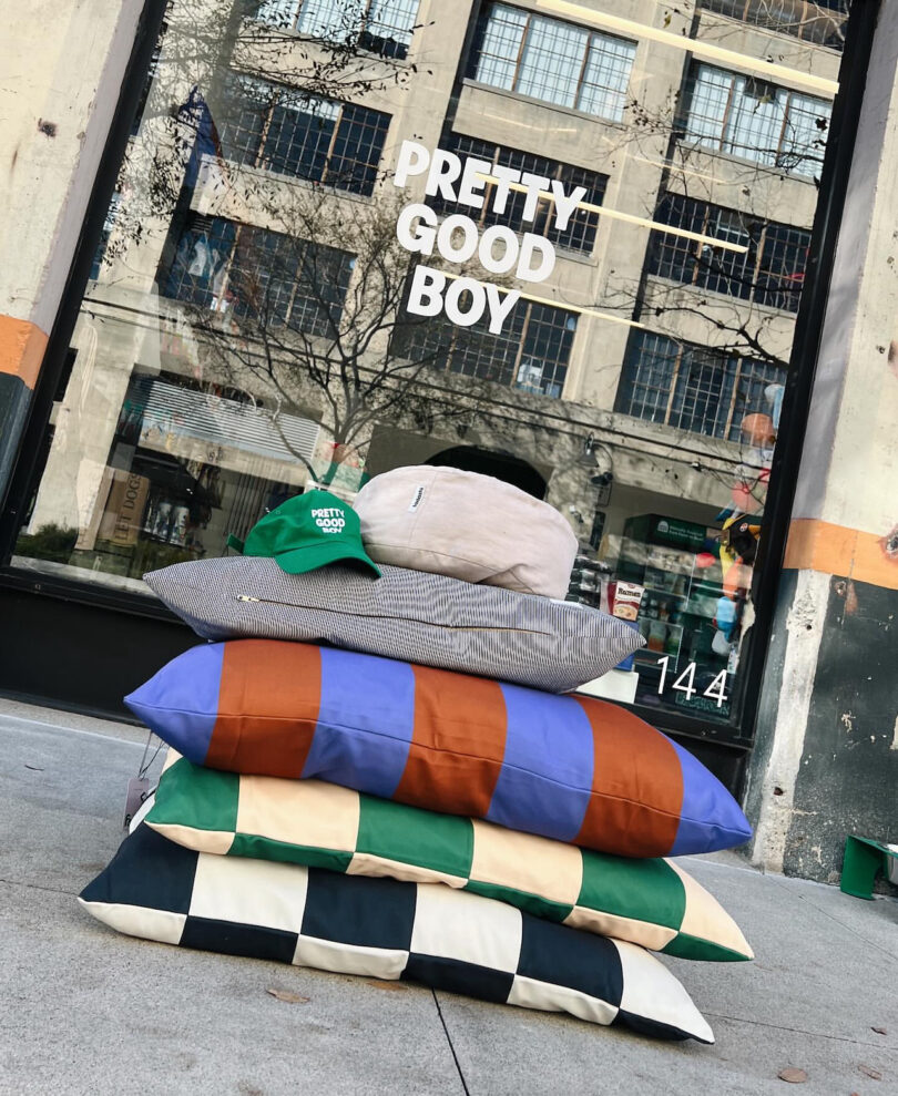 A stack of four striped and checkered cushions, two hats on top, placed on the sidewalk in front of a glass door with "PRETTY GOOD BOY" written on it