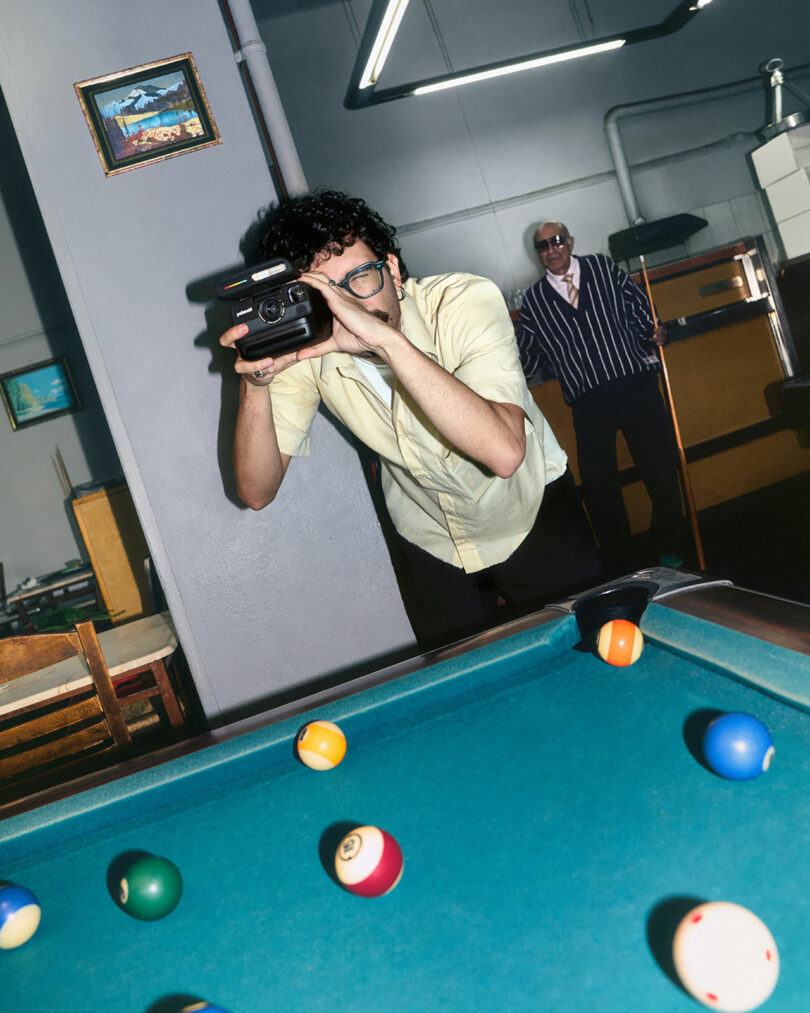 A man in glasses takes a photo with a camera beside a pool table, while another man in a striped jacket stands in the background.