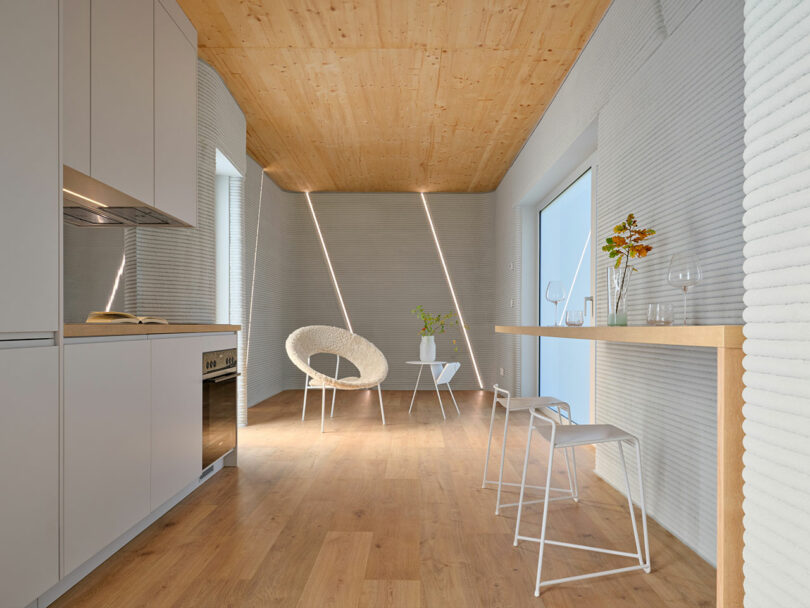 Modern minimalist kitchen and dining area with wood floors, light gray walls, built-in cabinets, a wall-mounted table, two white stools, and simple contemporary furniture.