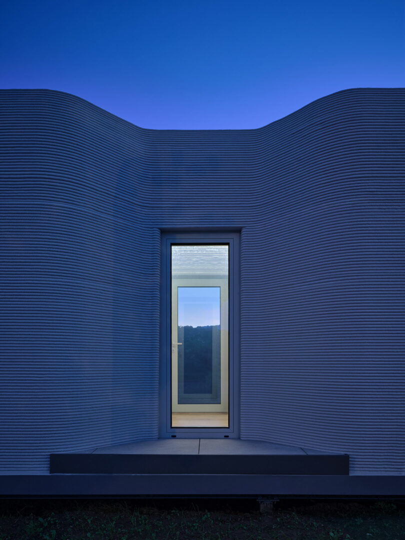 A modern building with smooth, wavy textured walls and a single illuminated door centered in the facade, photographed at dusk with a clear blue sky.
