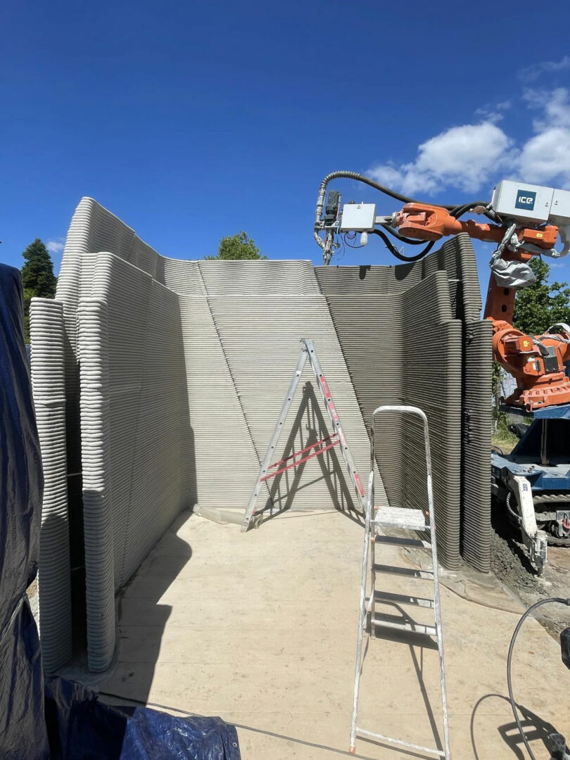 A robotic arm is 3D printing concrete walls of a structure on a construction site. Two ladders are positioned inside the unfinished building.
