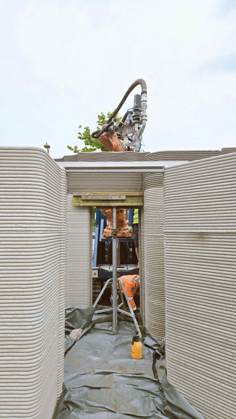 A 3D printer extrudes concrete to build the walls of a structure, with unfinished openings and construction materials visible on site.