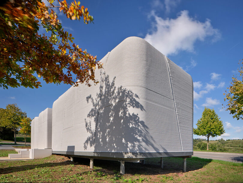 White, modern building with ribbed exterior panels elevated on stilts, casting shadows from nearby trees under a clear blue sky.
