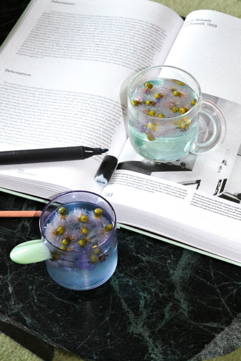 Two glass mugs with floral tea and ice cubes are placed on an open book, with a pen resting on the pages, atop a dark marble table