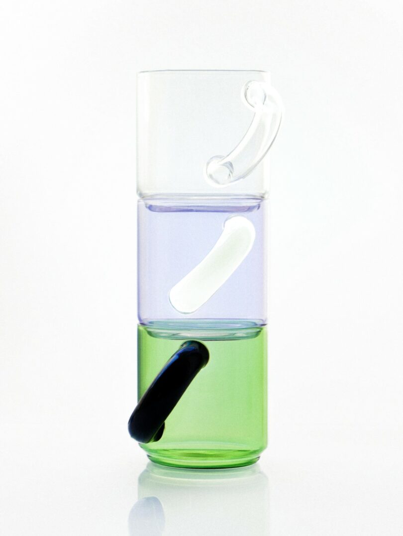 Three stacked glass cups, each with a curved handle in white, clear, and black, featuring transparent green, purple, and clear bodies, against a white background