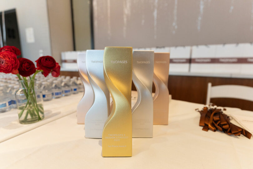 Metallic TWOPAGES awards labeled "IX DESIGN CONTEST" are displayed on a table adorned with red flowers, water bottles, and ribbons in the background.