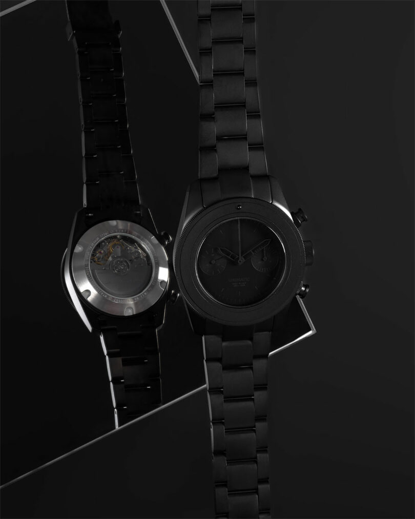 Black metal wristwatch displayed with its face and transparent case back visible, reflecting on a mirrored surface against a dark background.