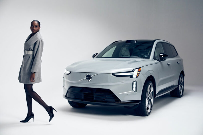 A woman in a gray coat and heels walks past a silver Volvo SUV in a minimalist studio setting.