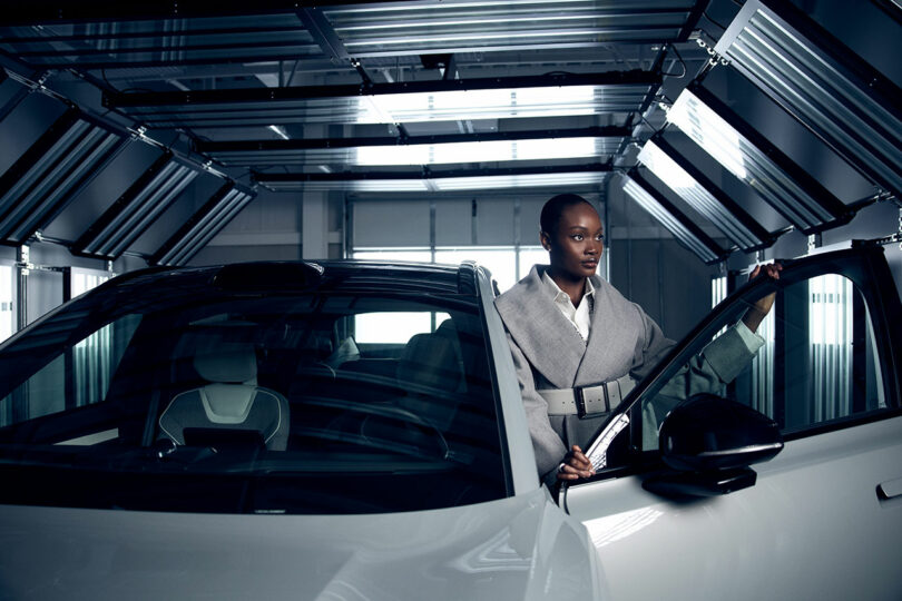 A woman in a gray coat stands by an open car door inside a modern, metallic garage.