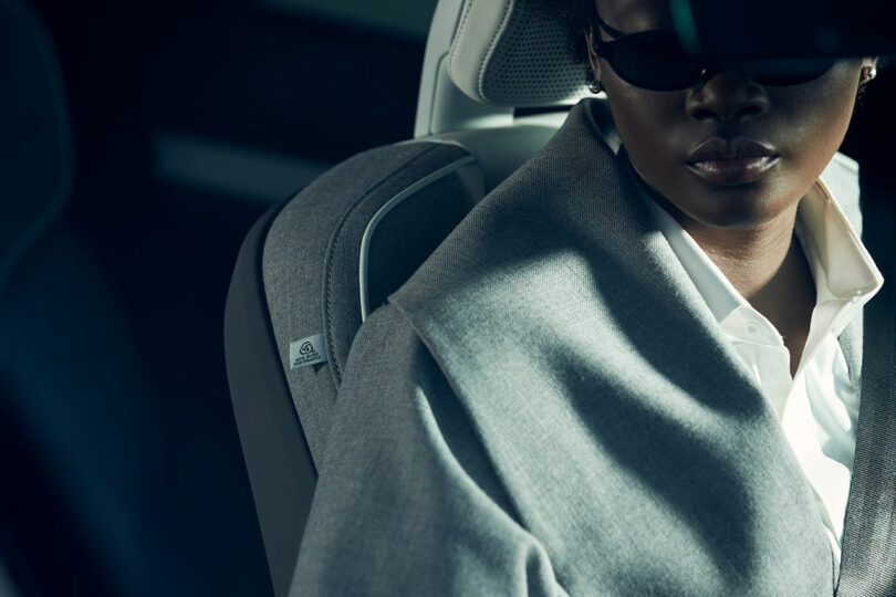 A person wearing a grey coat and sunglasses sits in a modern car seat, partially in shadow, with a close-up focus on the seat’s fabric and stitching.