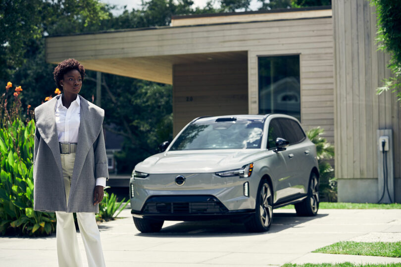 A person stands in front of a modern house with a silver electric SUV parked in the driveway, connected to a charging station.