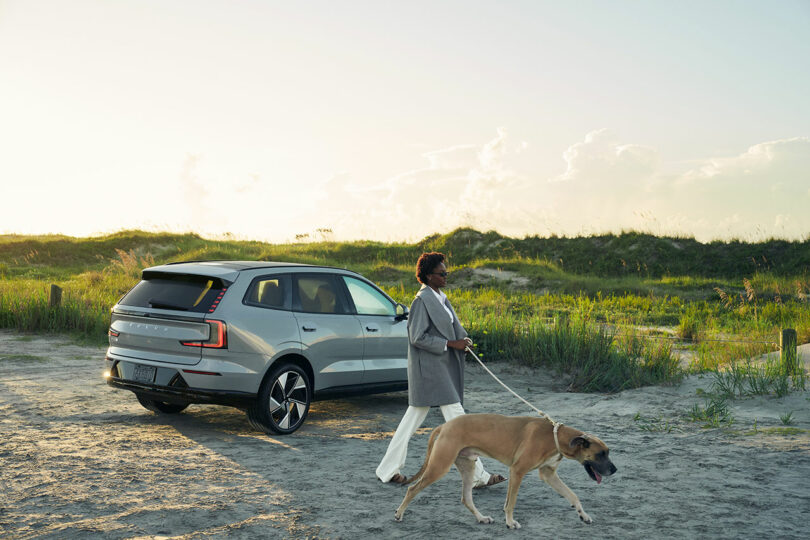 A person walks a large dog on a leash near a silver SUV parked on a sandy area with grassy dunes in the background.
