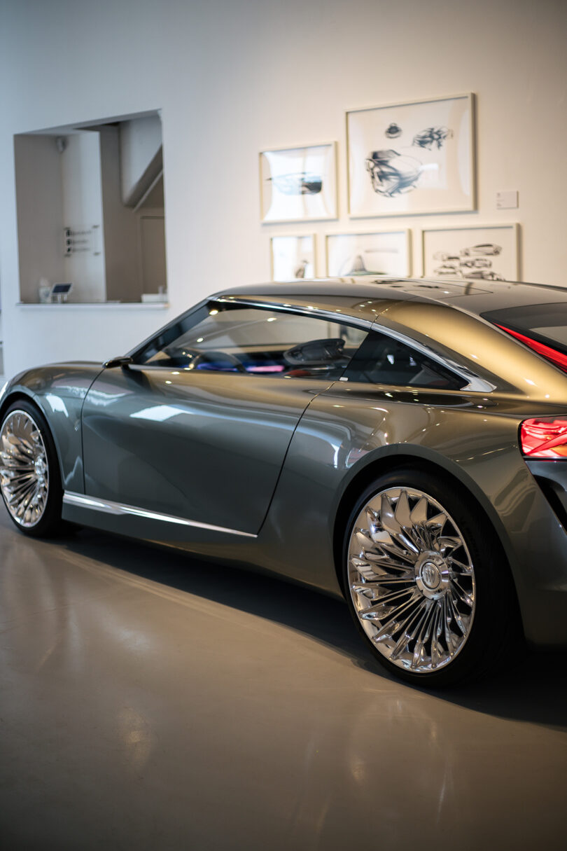 A sleek, modern silver concept car is parked indoors in a gallery space with framed art on the wall in the background.
