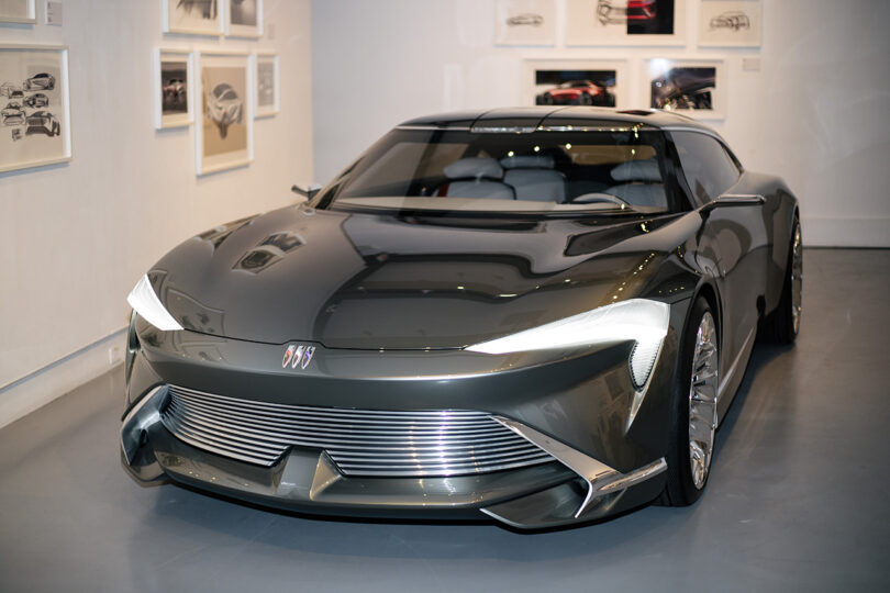 A sleek, dark gray concept car with futuristic design elements is displayed indoors, surrounded by framed automotive sketches on white walls.