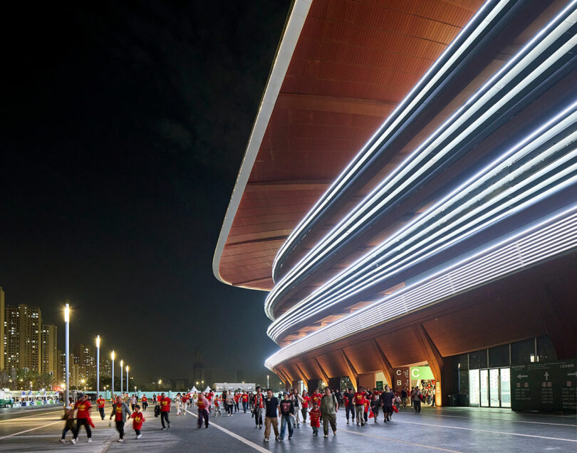 Crowds gather outside a modern, illuminated stadium at night, with city buildings visible in the background under a dark sky.
