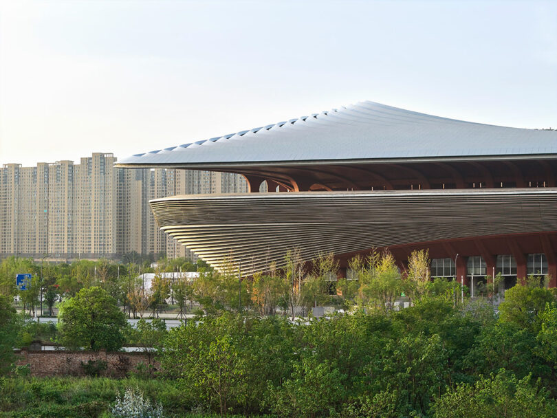 A modern stadium with a large, curved roof and layered exterior sits beside high-rise apartment buildings and greenery.