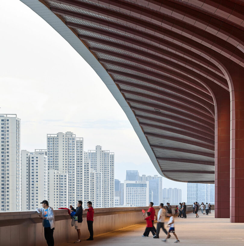 People stand on a terrace under a large, modern architectural overhang, with high-rise buildings visible in the background.