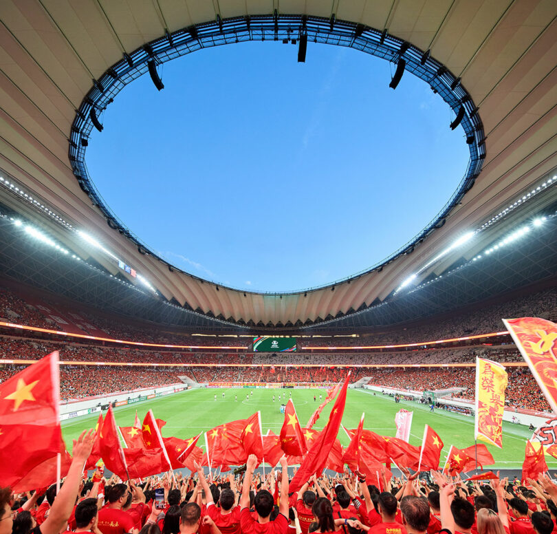 A large crowd waves Chinese flags in a packed modern stadium during a sports event, with a clear blue sky visible through the open roof.